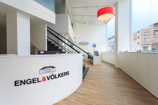 Interior de l'oficina d'Engel & Völkers. Recepció blanca amb logotip, escales negres, terra de fusta i llum penjant vermella.