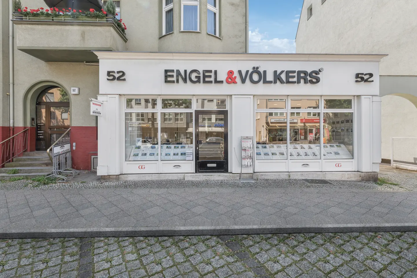 Engel & Völkers real estate office front with large windows displaying property listings. The building number 52 is visible on both sides.