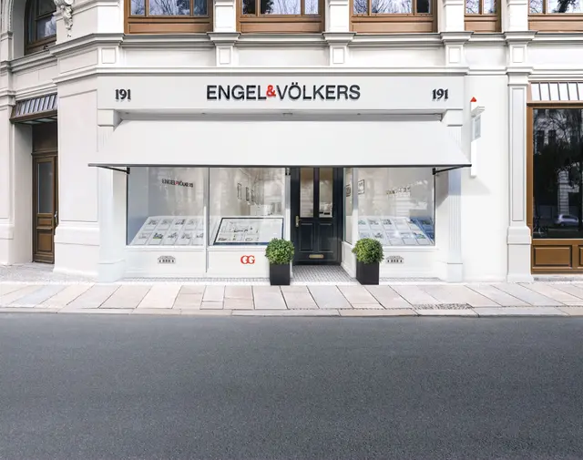 Engel & Völkers storefront at 191, with a white awning, large windows displaying property listings, and potted plants flanking the entrance.