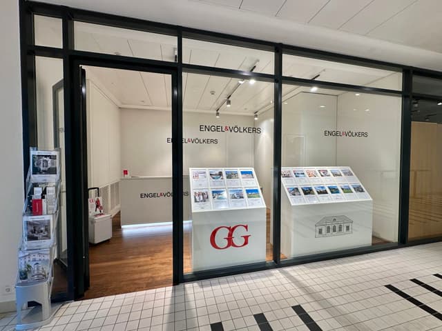 Ufficio immobiliare Engel & Völkers con finestre e porta in vetro con cornice nera. Display bianchi mostrano annunci immobiliari. Pavimento piastrellato.