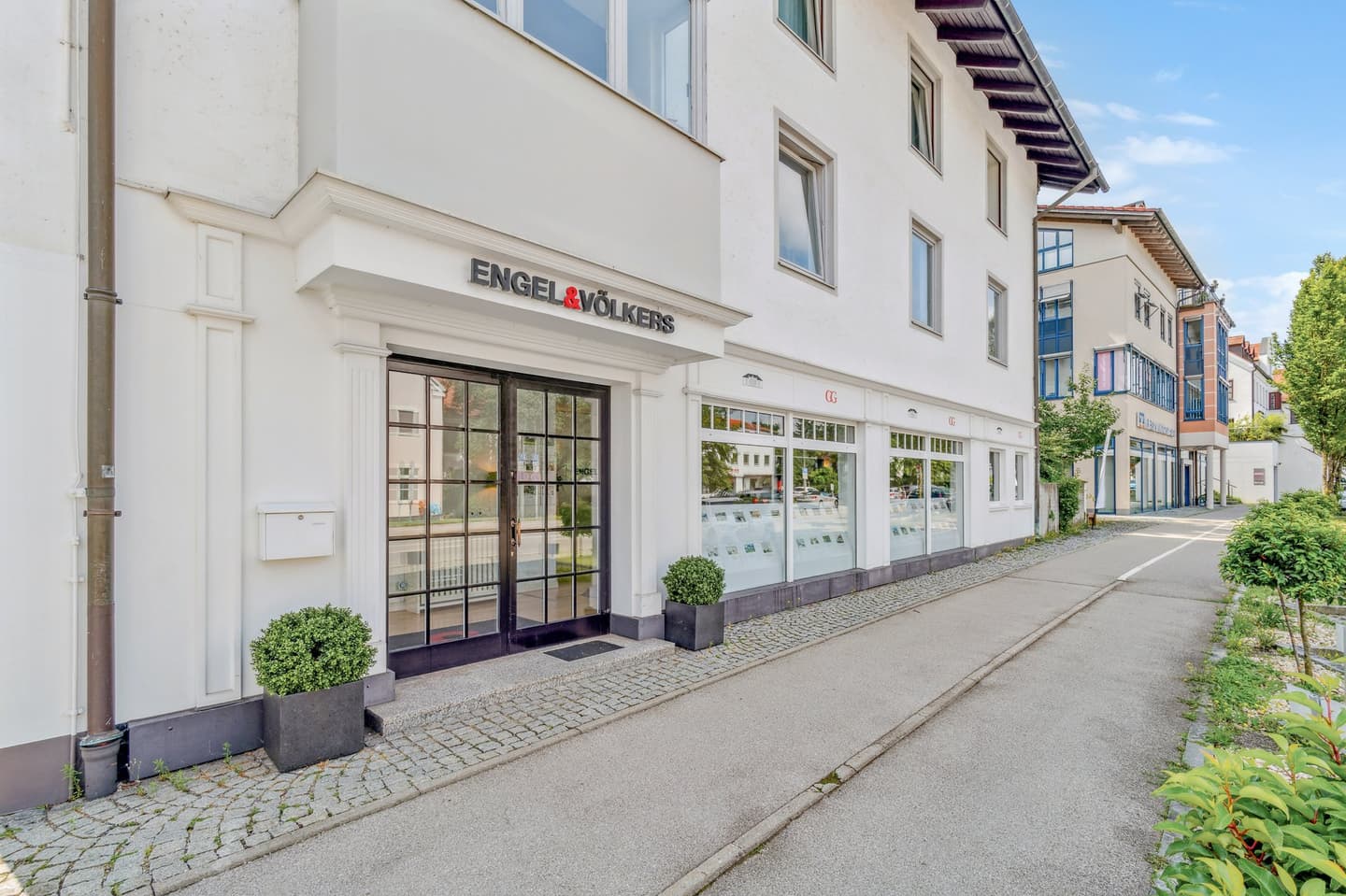 Engel & Völkers real estate office with white facade, black framed windows, and property displays. Two potted plants flank the entrance.