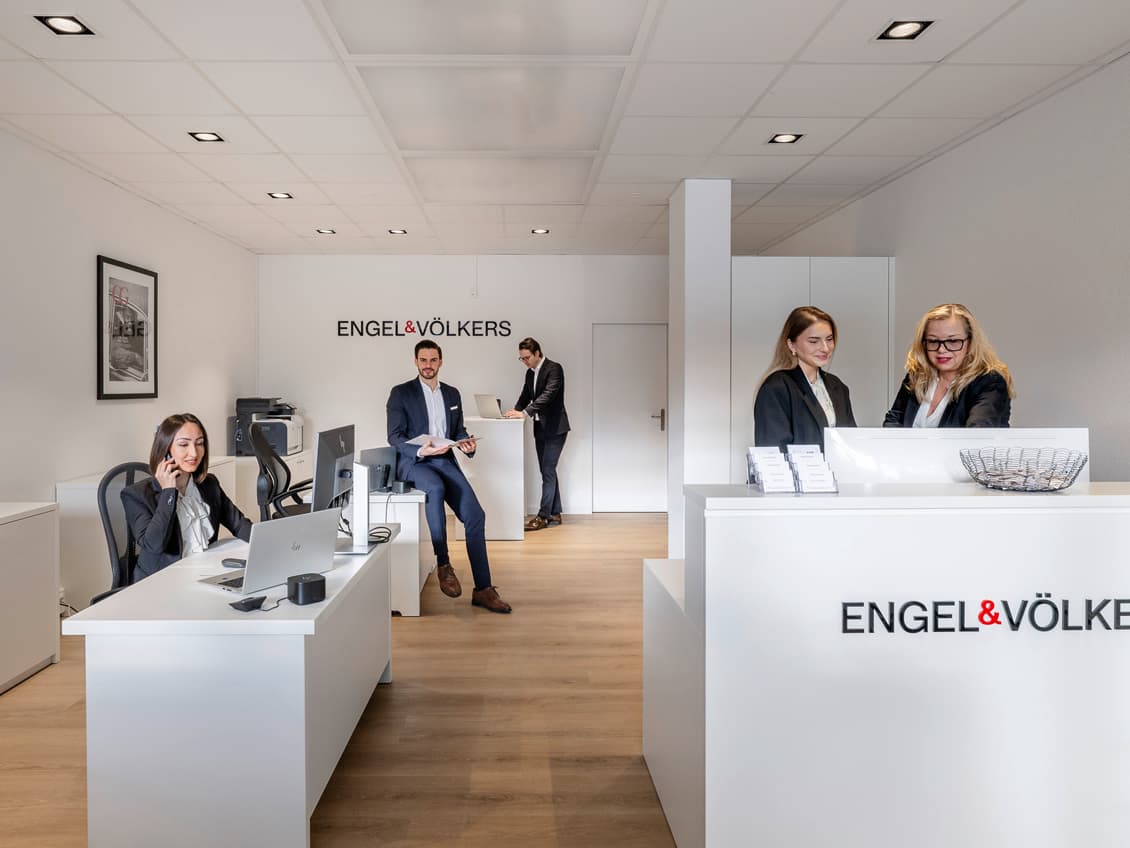 Engel & Völkers Immobilienbüro, Außenansicht. Ein roter Stuhl mit dem Firmenlogo steht vor dem Eingang. Immobilienangebote werden im Fenster ausgestellt.