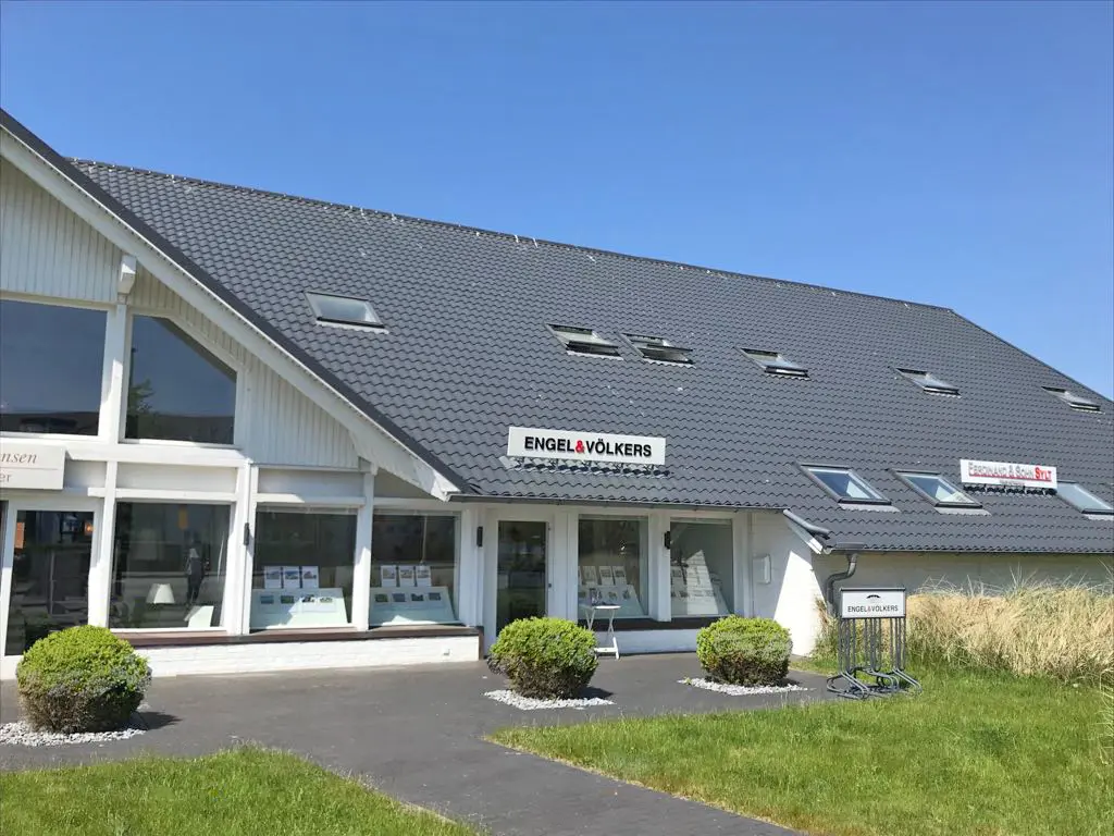Engel & Völkers real estate office with a gray tiled roof, white trim, and large windows displaying property listings. Tall ornamental grass in the foreground.